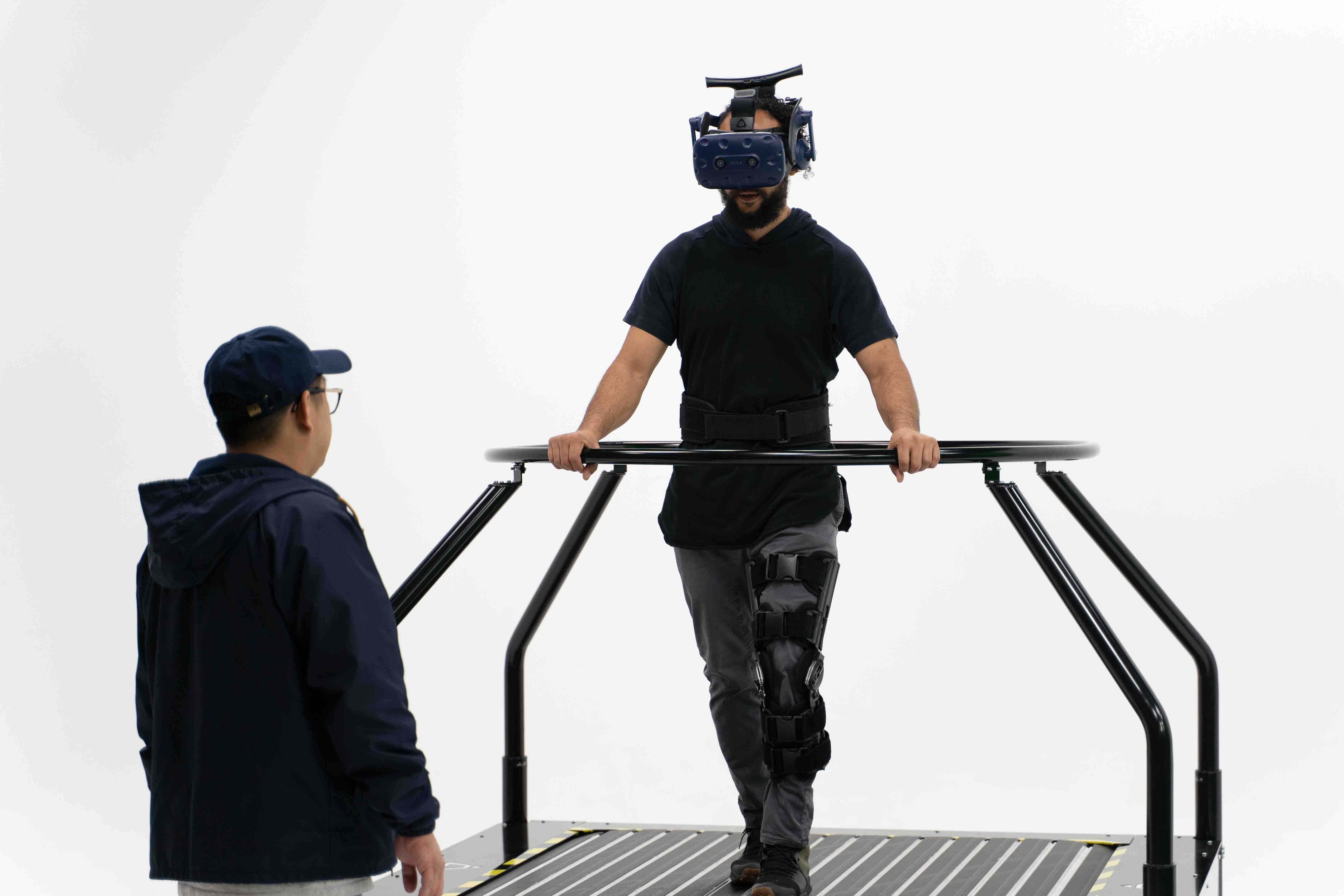 Man walking on an Infinadeck with a VR headset on, holding onto the rail, while another man watches